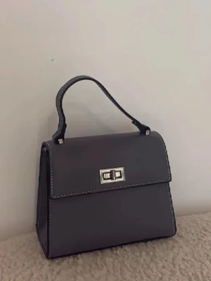 Grå handväska med silverdetalj - Stilren grå handväska i boxig form med kort handtag och silverfärgat vridlås framtill. Väskan har vita kontrastsömmar och är tillverkad i slätt syntetmaterial. Perfekt för dig som gillar minimalistisk och klassisk design. Aldrig använd 💕