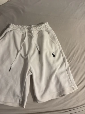 Vita shorts från Polo Ralph Lauren - Säljer ett par vita shorts från Polo Ralph Lauren med elastisk midja och snörning. Shortsen har två sidofickor och den klassiska broderade loggan i marinblått på vänster ben. Perfekta för sommaren och riktigt sköna tack vare mjukt bomullsmaterial.