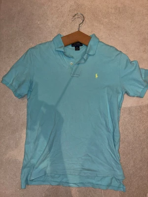 Ljusblå pikétröja från Ralph Lauren - Klassisk ljusblå pikétröja från Ralph Lauren med korta ärmar och krage. Tröjan har två knappar vid halsen och den ikoniska broderade loggan i gult på bröstet. Perfekt för en avslappnad och stilren look.