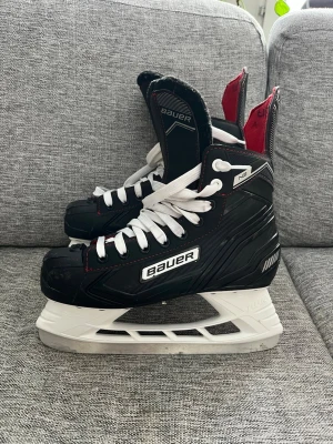 Svarta Bauer hockeyskridskor - Säljer ett par svarta hockeyskridskor från Bauer med vita snören och detaljer i rött och vitt. 