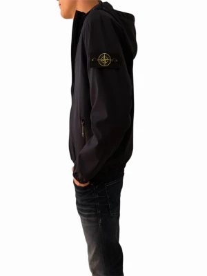 Mörkblå vindjacka från Stone Island - PASSAR L. Snygg mörkblå vindjacka från Stone Island med huva och dragkedja framtill. Jackan har två sidofickor med dragkedja och den klassiska Stone Island-loggan på vänster ärm. Perfekt för vår och höst.