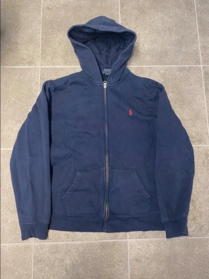 Ralph Lauren Zip Hoodie - Tröjan passar som Storlek S i herr. Mörkblå zip-hoodie från Polo Ralph Lauren med huva, dragkedja och fickor framtill. Klassisk röd Polo-logga på bröstet. Bekväm och stilren tröja i väldigt gott skick, perfekt för dig som gillar en clean, tidlös och sportig stil.