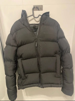 Svart dunjacka från Peak Performance - Säljer en svart dunjacka från Peak Performance med klassisk puffer-look. Jackan har huva, två stora framfickor och dragkedja framtill. Perfekt för kalla dagar och riktigt snygg till streetwear-stilen.