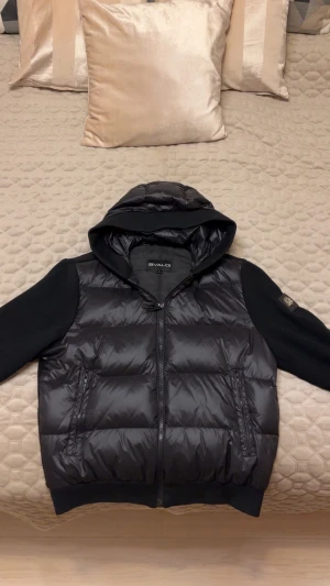 Svart cardigan med huva från Bvaldi - Svart cardigan från Bvaldi med snygg huva och ribbstickade ärmar. Framsidan är quiltad och glansig, medan baksidan och ärmarna är stickade. Jackan har dragkedja framtill och två fickor med dragkedja. Märkespatch på ärmen för extra stil. Inga defekter, nypris 2500kr hör av er med frågor passar mig perfekt och är 191 cm.