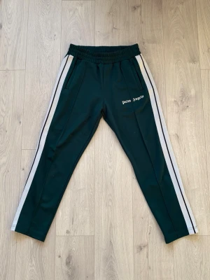 Palm Angels Track Pants - Mörkgröna byxor från Palm Angels i fint skick överlag. Tyvärr har storlekslappen rivits av men Certilogo finns kvar för äkthetsverifiering. En liten reva på höger ben vilket endast syns vid noga inspektion. 