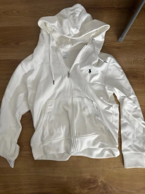 Vit hoodie från Polo Ralph Lauren - Snygg vit hoodie med dragkedja från Polo Ralph Lauren. Klassisk design med huva och dragsko, två stora fickor framtill och den ikoniska lilla loggan broderad på bröstet. Perfekt för en clean och avslappnad stil.