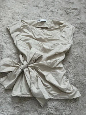 Beige omlottopp med knyt från Zara - Säljer en beige omlottopp från Zara i storlek M. Toppen har en cool asymmetrisk halsringning och knyt i midjan som ger en snygg siluett. Ärmlös design och mjukt bomullsmaterial gör den både bekväm och trendig. Perfekt att styla med jeans eller kjol🦢🤍