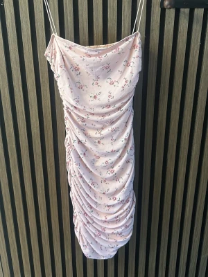 Ljusrosa blommig klänning NLY One - Supersöt ljusrosa kortklänning från NLY One med smala axelband och blommigt mönster. Klänningen har rynkade detaljer längs sidorna som ger en snygg draperad effekt. Perfekt för sommarens alla tillfällen!