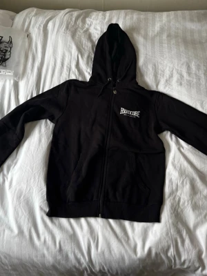  100%Hardcore.    ZIPUP - Säljer en svart hoodie med dragkedja från 100%Hardcore. Tryck med logga på bröstet och stort tryck på ryggen där det står '100% Hardcore Wear it with pride'. Hoodien har huva med dragsko och två fickor framtill. Perfekt för dig som gillar streetwear.