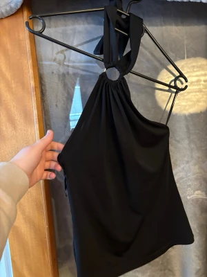 Svart oneshoulder halterneck topp - Snygg svart oneshoulder topp med halterneck och knyt i nacken. Toppen har en slät och enkel design som ger en stilren look. Perfekt för dig som vill ha något trendigt och bekvämt till sommaren.