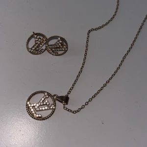  halsband och örhängen - Set med halsband och örhängen. Båda har rund hängande design med LV-logga i guldtonad metall och glittrande stenar. Kedjan är tunn och smidig, och loggorna är inramade av en tvinnad kant. Perfekt för dig som gillar lyxiga detaljer.