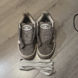 Grå Adidas Campus sneakers - Säljer ett par grå Adidas Campus sneakers med klassiska vita ränder på sidan och Adidas-logga på plösen. Skorna har rund tå, platt sula och är tillverkade i mocka med vita detaljer. Medföljer vita skosnören. Perfekt för dig som gillar retrostil och streetwear.