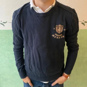 Mörkblå tröja från Morris med emblem - Snygg mörkblå tröja från Morris med broderat emblem och text på bröstet. Tröjan har rund halsringning, ribbade muddar och är tillverkad i mjuk bomull. Perfekt för dig som gillar klassisk och stilren look.