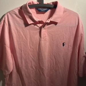 Rosa Polo Golf t-shirt från Ralph Lauren - Säljer en klassisk rosa t-shirt från Polo Golf Ralph Lauren med krage och knappslå. T-shirten har korta ärmar och den ikoniska broderade loggan på bröstet. Tillverkad i 100% bomull för en skön och luftig känsla. Perfekt för dig som gillar stilren design.