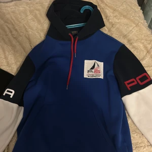 Blå hoodie Polo Ralph Lauren P-15 - Snygg blå hoodie från Polo Ralph Lauren med P-15 Offshore Racing patch på bröstet och amerikansk flagga på ryggen. Tröjan har kontrastfärgade ärmar i mörkblått och vitt, röda snören i huvan och tryckta detaljer på båda ärmarna. Perfekt för dig som gillar sportig och cool stil.