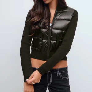 Svart croppad pufferjacka med stickade ärmar - Snygg svart croppad pufferjacka med glansig front och stickade långärmade sidor. Jackan har dragkedja framtill och två fickor. Perfekt för dig som gillar en modern och edgy stil.