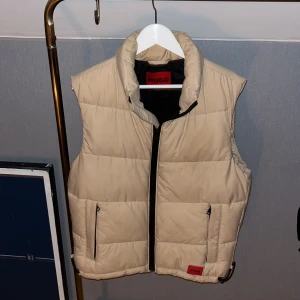 Beige dunväst från Hugo boss - Snygg beige dunväst från Hugo med svart innerfoder och markerad röd logotyp på insidan. Västen har hög krage, två fickor med dragkedja och quiltad design. Perfekt för lager-på-lager och chill stil under kyliga dagar.
