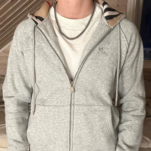 Grå hoodie från Burberry med huva - Säljer en grå hoodie från Burberry i storlek M. Tröjan har dragkedja, snörning vid huvan och klassisk Burberry-logga broderad på bröstet. Insidan av huvan har ett snyggt randigt mönster i beige, svart och rött. Perfekt för dig som gillar stilrena märkesplagg.