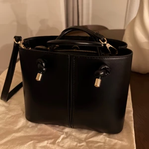 Svart handväska med gulddetaljer - Stilren svart handväska i skinn med både handtag och axelrem. Väskan har guldfärgade metalldetaljer och dragkedja, samt snygga knyt-detaljer framtill. Insidan har flera fack för smart organisering. Perfekt storlek för att få med allt viktigt. Väskan är från Zara. 