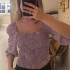 Ljuslila blus med puffärmar - Superfin ljuslila blus med smock och puffiga trekvartsärmar. Blusen har en fyrkantig urringning och volangdetaljer vid hals och ärmslut💜