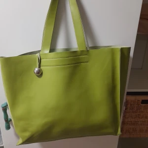 Grön shoppingväska från Furla - Stor och stilren shoppingväska från Furla i äkta skinn, tillverkad i Italien. Väskan är ljusgrön med raka linjer och har två breda handtag. Utsidan är slät och pryds av en silverfärgad rund detalj. Perfekt för dig som gillar minimalistisk design och vill sticka ut.