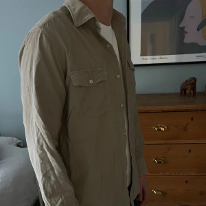 Beige manchester overshirt med fickor - Snygg beige overshirt i manchestertyg med klassisk krage och två bröstfickor med knappar. Skjortan har långa ärmar och knäppning framtill. Perfekt att ha över en t-shirt. Enkel och stilren modell som funkar till jeans.