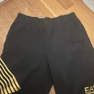 Svarta EA7 shorts med guldtryck - Svarta shorts från EA7 Emporio Armani med elastisk midja och snygga guldiga detaljer. Ena benet har guldtryck med logga och det andra har coola guldiga ränder. Mjukt material som känns skönt mot huden, perfekt för chill eller sport.