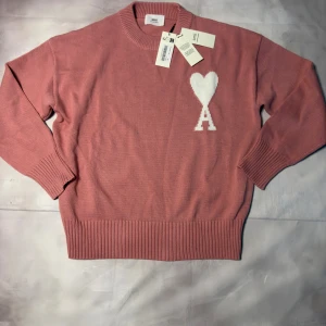 Rosa stickad tröja från Ami Paris - Snygg rosa stickad tröja från Ami Paris med vit hjärtlogga och bokstaven A på bröstet. Tröjan har rund hals, ribbade muddar och är tillverkad i mjuk bomull. Perfekt för dig som gillar stilrena detaljer och vill ha en trendig look.