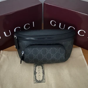 Svart magväska  med mönster - Snygg svart magväska från med klassiskt mönster och två praktiska fack med dragkedja. Perfekt storlek för det viktigaste och stilren design som lyfter din outfit. Väskan har detaljer i skinn och silverfärgade dragkedjor.