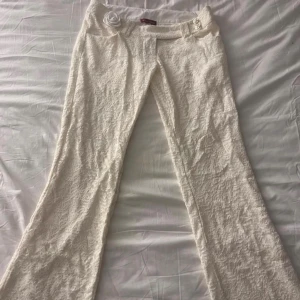 Vita spetsbyxor med blomdetalj S - Unika vita byxor från L'ange Vivant i storlek S, täckta av spets med en snygg rosdetalj framtill. Byxorna har bootcut-modell och dekorativa knappar i midjan. 