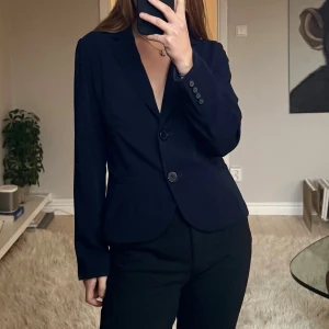 Mörkblå kavaj från Gerry Weber - Snygg mörkblå kavaj från Gerry Weber med klassisk krage och två knappar framtill. Kavajen har figurnära passform, långa ärmar med knappdetaljer och två diskreta fickor. Perfekt för dig som gillar stilrena och tidlösa plagg.