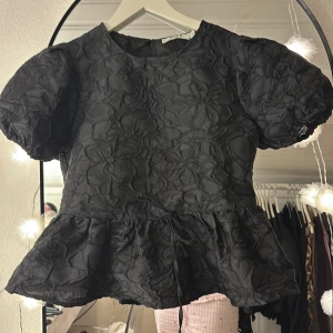 Svart blus med puffärm och volang från nakd  - Säljer en svart blus från NA-KD med puffiga korta ärmar och volang nedtill. Blusen har ett quiltat blommigt mönster och rund halsringning. Materialet är glansigt och ger en lyxig känsla. Perfekt för dig som gillar statement-plagg med cool struktur. Använd en gång