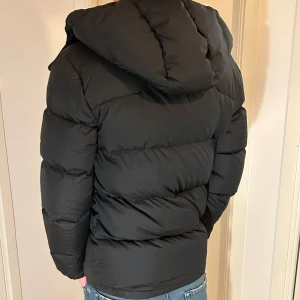 Svart Ralph Lauren jacka  - Svart pufferjacka från Polo Ralph Lauren med röd logga på bröstet. Jackan har huva, dragkedja framtill och två sidofickor med dragkedja. Den är vadderad och har en clean, modern look som passar till jeans och hoodie. Jackan är i nyskick. Endast använd 2ggr. Passar bra om du är 178cm. 
