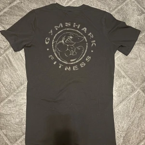 Svart Gymshark t-shirt med tryck - Svart t-shirt från Gymshark med stort tryck av en muskulös haj och texten 'GYMSHARK FITNESS' på ryggen. Framtill finns en mindre logga på bröstet. T-shirten har rund hals och korta ärmar, perfekt för gymmet eller chill. Materialet är mjuk bomull.