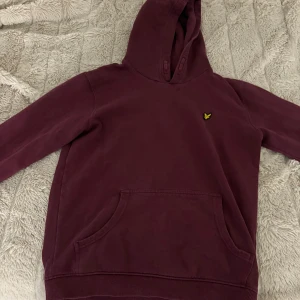 Vinröd hoodie från Lyle & Scott - Säljer en vinröd hoodie från Lyle & Scott Junior med klassisk gul logga på bröstet. Tröjan har huva, magficka och långa ärmar. Mjuk bomullskvalitet och ribbade muddar vid ärmslut och nederkant.