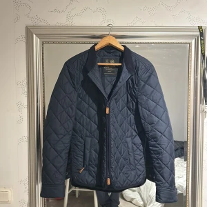 Mörkblå quiltad jacka från Morris - Quiltad jacka från Morris i mörkblå färg med snyggt rutmönster och broderad logga på bröstet. Jackan har dragkedja framtill, klassisk krage och två fickor med detaljer i brunt. Perfekt för dig som gillar stilren och tidlös design.