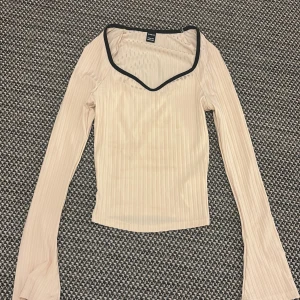 Beige ribbad långärmad topp Shein - ( mindre i storleken och ganska tajt)Säljer en beige ribbad långärmad topp från Shein i storlek XXS. Toppen har en snygg v-ringad hals med svart kant och är figurnära i modellen. Materialet är stretchigt och lätt, perfekt för att styla med jeans eller kjol.