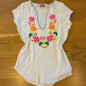 Vit blus med broderade blommor Stella Forest - Vit blus från Stella Forest med färgglada broderade blommor framtill. Djup v-ringning med lila kant och korta ärmar. Perfekt för en somrig look med unika detaljer och mjukt bomullsmaterial.