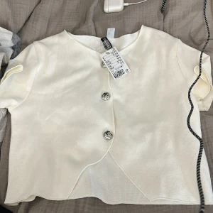 Vit omlott blus med knappar från h&m - Säljer en vit blus från h&m med snygg omlott-design och tre dekorativa knappar framtill. Blusen har korta ärmar och är tillverkad i ett mjukt stickat material. Perfekt för dig som gillar stilrena och trendiga toppar.