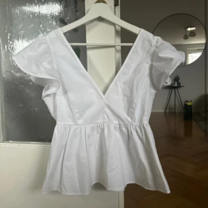 Vit blus med volangärm  -  Blusen har djup v-ringning både fram och bak, korta volangärmar och en peplumliknande nederdel som ger en snygg siluett. Ny med prislapp!!