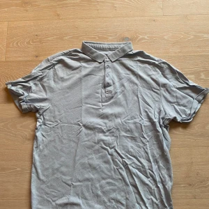 Grå piké från Clean Cut Copenhagen XXL - Snygg grå pikétröja från Clean Cut Copenhagen i storlek XXL. Tröjan har klassisk krage, korta ärmar och tre knappar framtill. Materialet är bomull med en cool, strukturerad yta och ett litet broderat X på bröstet. Perfekt för en chill och stilren look.