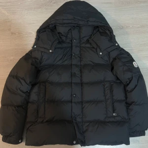 Moncler Vezere - Svart Moncler jacka med  huva, quiltad design och logotyp på ärmen. Jackan har både dragkedja och knappar framtill, samt praktiska fickor. Perfekt för kalla dagar och har en riktigt schysst puffer-look.
