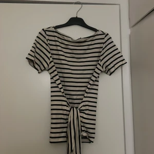 Randig topp med knytband - Säljer en vit och svart randig topp med korta ärmar och bred halsringning. Toppen har långa knytband i sidorna som kan knytas i midjan för en snygg siluett. Materialet känns mjukt och stretchigt, perfekt för en chill och trendig look.