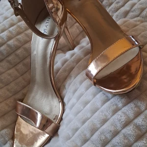 Metallic roséguldiga klackpumps NLY - Snygga pumps i metallic roséguld från NLY Shoes med smal hög klack och öppen tå. Skorna har en enkel rem över tårna och justerbart spänne vid ankeln. Materialet är syntet med glansig finish som ger en lyxig vibe. Perfekta för att sticka ut med en cool och trendig look.