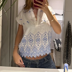 Vit blus med blå broderi och v-ringning - Säljer en söt vit blus med blå broderade detaljer och v-ringning. Blusen är croppad med korta ärmar och har ett unikt mönster längs nederkanten. Materialet känns lätt och luftigt, perfekt för varma dagar. 