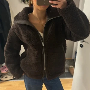Mörkbrun teddyjacka med dragkedja - Mysig mörkbrun teddyjacka med hög krage och tvåvägsdragkedja framtill. Jackan har en loose passform och är tillverkad i fluffigt fleece-material som ger en skön och avslappnad vibe. Perfekt för kyliga dagar och enkel att styla med jeans eller hoodies.