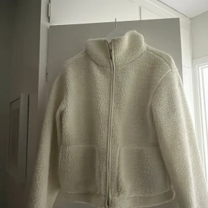 Vit teddyjacka med dragkedja - Mysig vit teddyjacka med hög krage och hel dragkedja framtill. Jackan har två stora fickor och är tillverkad i fluffigt fleece-material som håller dig varm och skön. Perfekt för kyliga dagar och enkel att matcha med din vardagsstil.