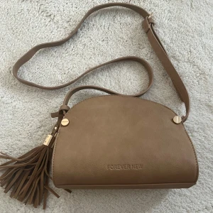Brun axelväska med fransar Forever New - Snygg brun axelväska från Forever New i skinnimitation med justerbar rem och coola guldfärgade detaljer. Väskan har en halvmåneformad design och en stor fransdetalj på sidan som ger extra stilpoäng. Perfekt storlek för det viktigaste och enkel att matcha med din outfit.