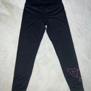 Svarta leggings från Röhnisch S - Svarta leggings från Röhnisch i storlek S med hög midja och rosa detaljer på insidan samt ett rosa tryck på ena benet. Tillverkade i stretchigt syntetmaterial som passar perfekt för träning eller sportiga aktiviteter.