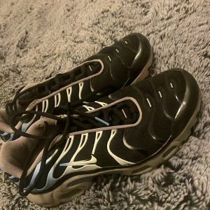 Nike Air Max Plus svart/vit EUR 40 - Nike Air Max Plus sneakers i svart med vita och ljusblå detaljer. Snygg vågig design på ovandelen, meshmaterial för bra ventilation och robust gummisula med TN-logga undertill. Skorna har snörning och en rund tå. Perfekt för dig som gillar sportig och modern stil.
